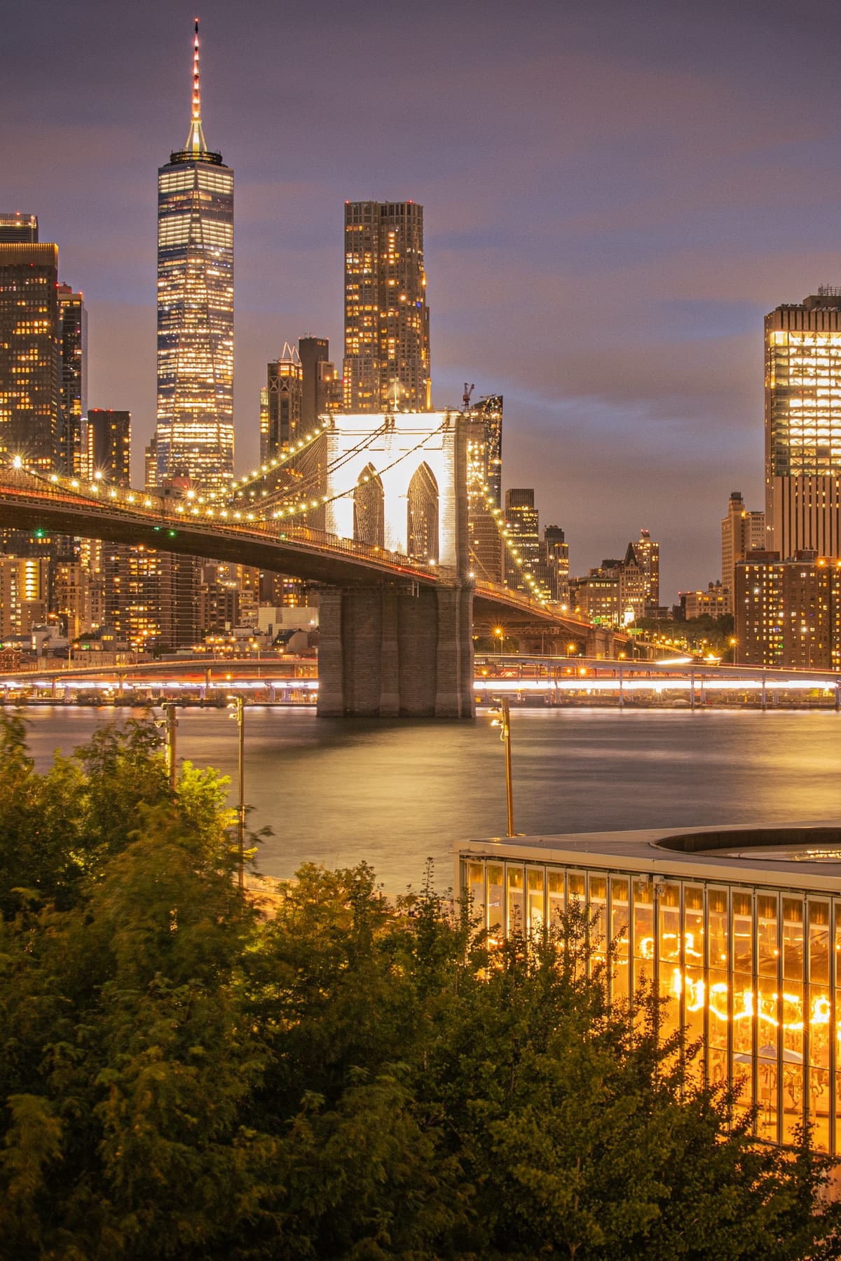 Brooklyn Bridge and the Lower Manhattan skyline at night, with the lit carousel pavilion on the waterfront below.